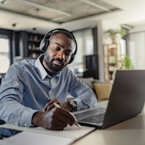 Man with headphones writing beside laptop.
