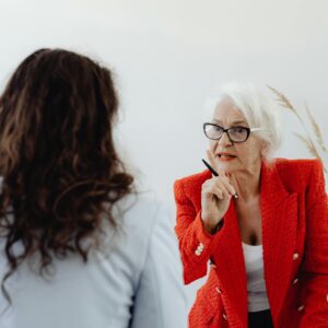 Two women engaged in a serious conversation, one in a red blazer gesturing thoughtfully.