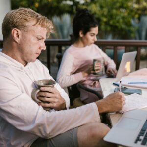 Two professionals working outdoors, one with coffee and notes, the other on a laptop.