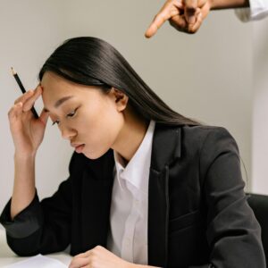 Stressed woman in business attire holding a pen, with a hand pointing at her.