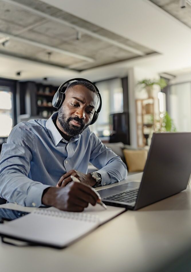 Man with headphones writing beside laptop.