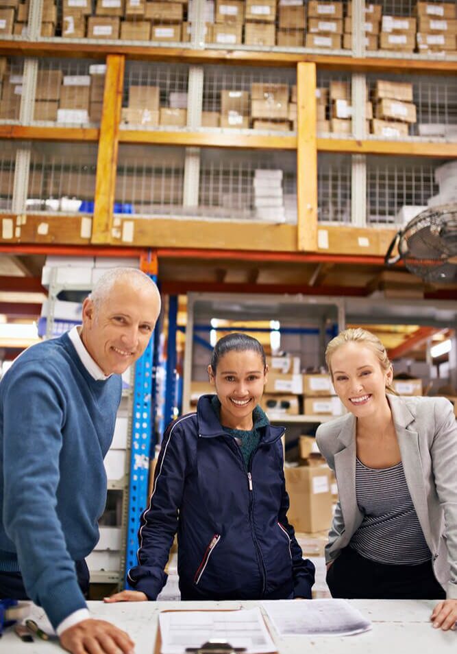 Three professionals smiling in a warehouse with shelves of boxes.