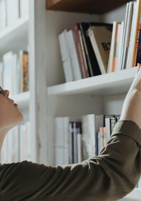 A woman reaching for a book on a white bookshelf.