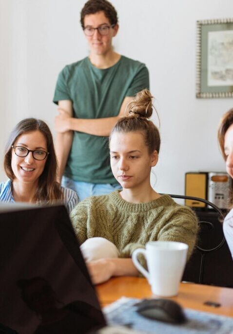 Group of young adults and a woman gathered around a laptop, engaged and smiling.