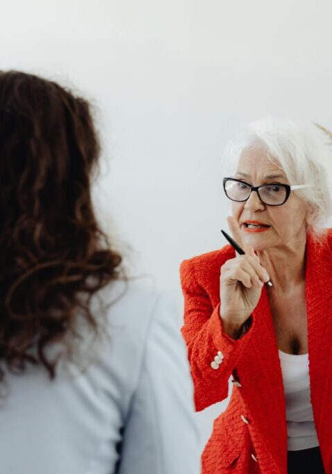 Two women engaged in a serious conversation, one in a red blazer gesturing thoughtfully.