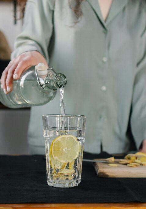 A person pours water into a glass with lemon slices in a cozy kitchen.
