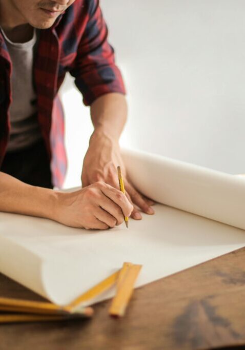 A person sketching on rolled blueprint paper at a wooden table.