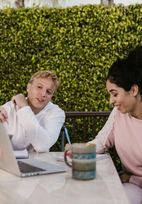 Two women engaged in a conversation at an outdoor table with a laptop and coffee.
