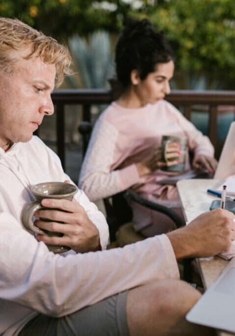 Two professionals working outdoors, one with coffee and notes, the other on a laptop.