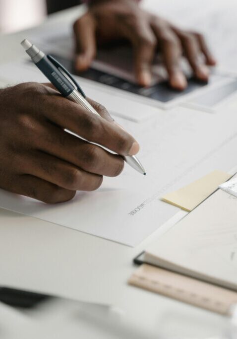 Person writing on paper with a pen at a cluttered desk.