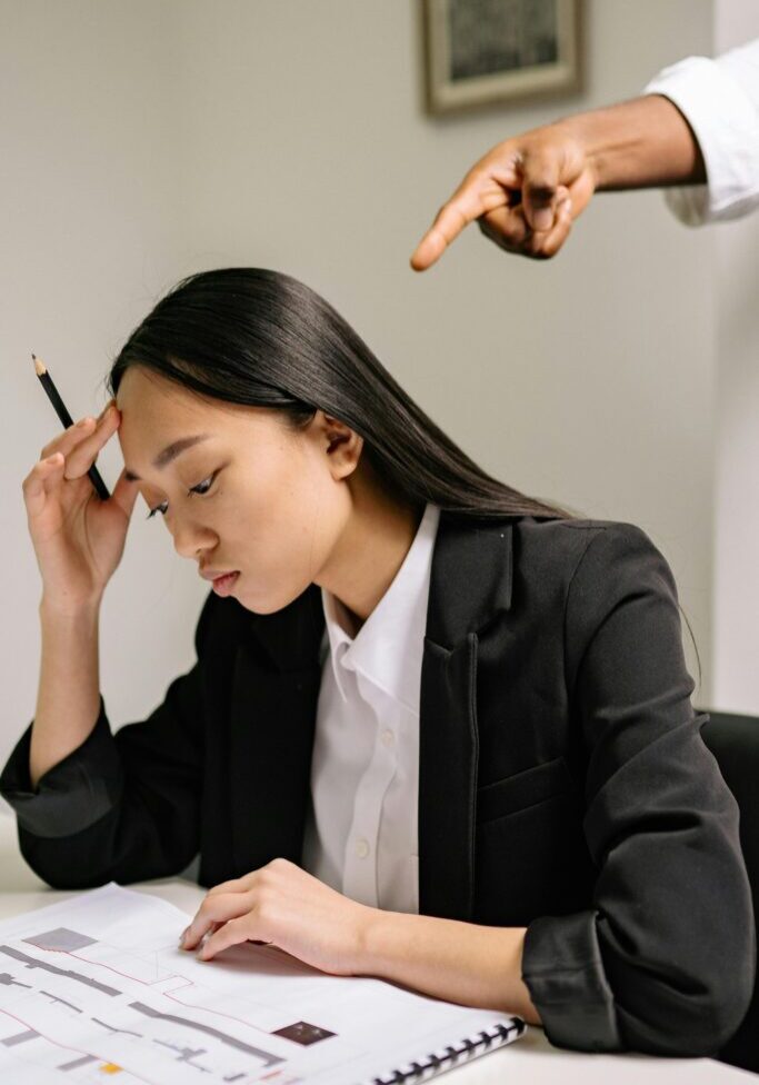 Stressed woman in business attire holding a pen, with a hand pointing at her.
