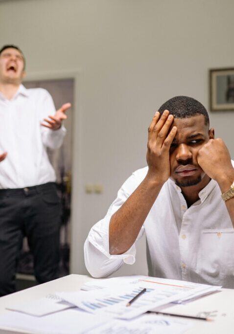 Stressed man at desk while another argues in background.